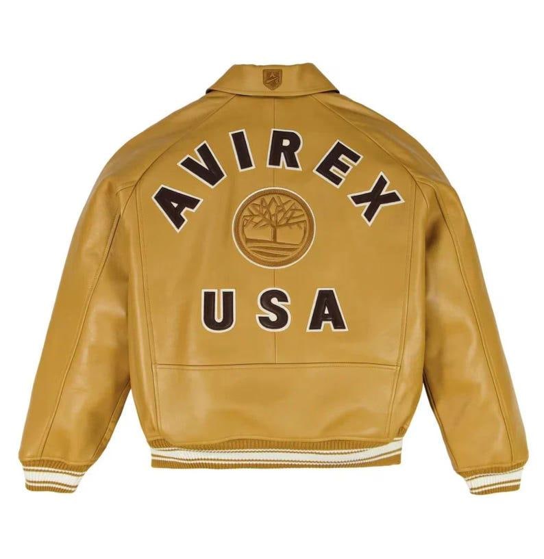 May include: A tan leather jacket featuring a classic collar and ribbed cuffs and hem with white stripes. The back displays the text AVIREX in a dark brown arch, a circular tree logo emblem in the center, and the text USA in dark brown letters below.