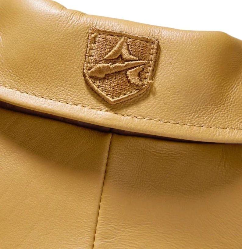 May include: A tan leather garment featuring a folded collar and a centered, embroidered shield-shaped patch. The patch displays a stylized letter A symbol in a matching tan thread, with visible stitching along the edges of the leather collar.