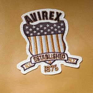 May include: An embroidered patch on a tan leather surface featuring the text AVIREX at the top, a shield with white stars on a dark brown background, vertical stripes, and a banner reading ESTABLISHED NY 1975 in dark brown and white thread.