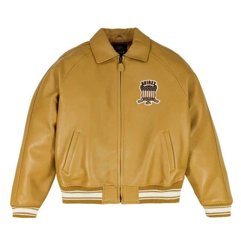 May include: A tan leather jacket featuring a full-length front zipper, a classic collar, and ribbed cuffs and hem with white stripes. On the left chest, there is an embroidered patch displaying the text AVIREX above a shield with a flag design.