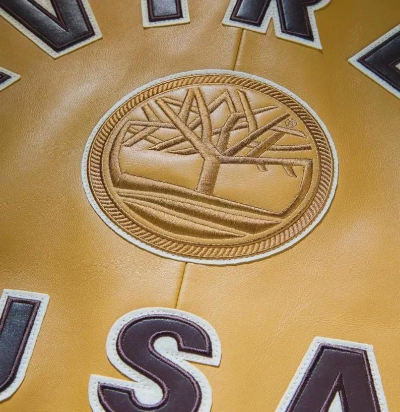 May include: A tan leather jacket featuring a large circular embroidered tree logo in the center. The design includes dark brown and white block lettering that spells out USA and other partial text around the central emblem on the smooth surface.