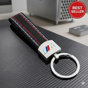 May include: Black leather key fob with red, white, and blue stitching. The fob features a silver-colored metal plate with the BMW logo and a silver key ring. A red circle with "BEST SELLER" is in the background.