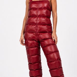 Pode incluir: Macacão de neve acolchoado vermelho com alças largas. A peça única apresenta um design fofo e acolchoado, cobrindo o torso e as pernas. O macacão de neve é de cor vermelha vibrante, adequado para atividades em climas frios.