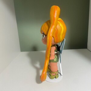 Op de afbeelding: Een beeldje van een cartoonfee met geel en oranje haar in een paardenstaart. De fee draagt een groene outfit en heeft blauwe vleugels. Het beeldje staat op een witte basis.