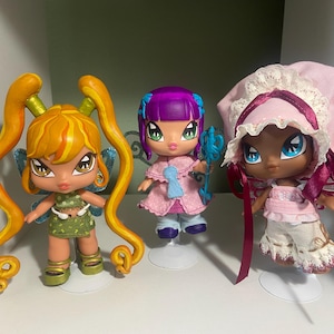 Op de afbeelding: Drie Bratz-achtige poppen met grote ogen en gestileerde kenmerken. De poppen hebben verschillende haarkleuren en outfits. E&eacute;n pop heeft oranje haar in staartjes en een groene jurk. Een andere heeft paars haar en een roze jurk. De derde pop draagt een roze muts.