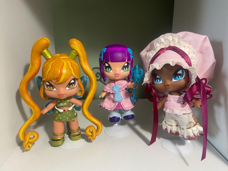 Op de afbeelding: Drie Bratz-achtige poppen met grote ogen en gestileerde kenmerken. De poppen hebben verschillende haarkleuren en outfits. E&eacute;n pop heeft oranje haar in staartjes en een groene jurk. Een andere heeft paars haar en een roze jurk. De derde pop draagt een roze muts.