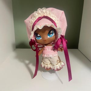 May include: A doll with dark skin, large blue eyes, and a pink bonnet with lace trim and ribbon. The doll wears a pink top and cream-colored shorts with a ruffled hem. The doll is standing on a clear stand.