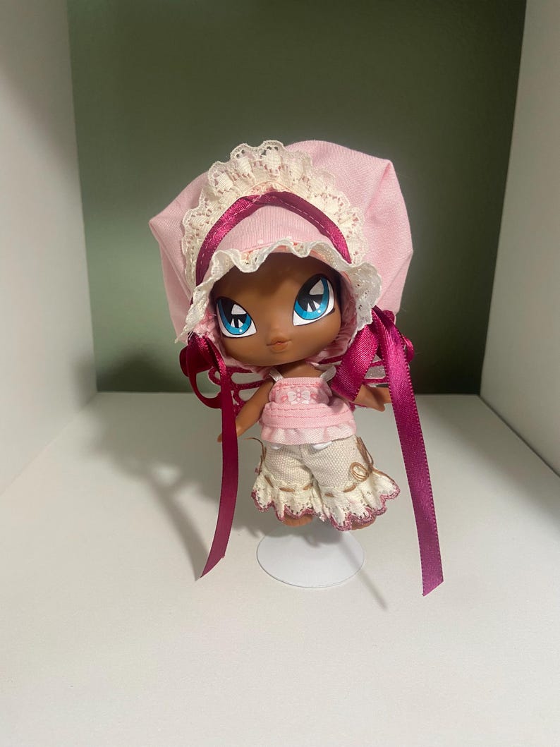 May include: A doll with dark skin, large blue eyes, and a pink bonnet with lace trim and ribbon. The doll wears a pink top and cream-colored shorts with a ruffled hem. The doll is standing on a clear stand.