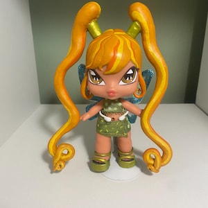 Op de afbeelding: Een pop met oranje haar in twee lange, krullende staarten. De pop heeft grote ogen, blauwe vleugels en draagt een groene outfit met een bloemenpatroon. De pop staat op een witte basis.