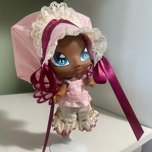 May include: A doll with dark skin, large blue eyes, and a pink bonnet with lace trim and burgundy ribbons. The doll wears a pink dress and cream-colored shorts with lace accents. It stands on a white metal stand.