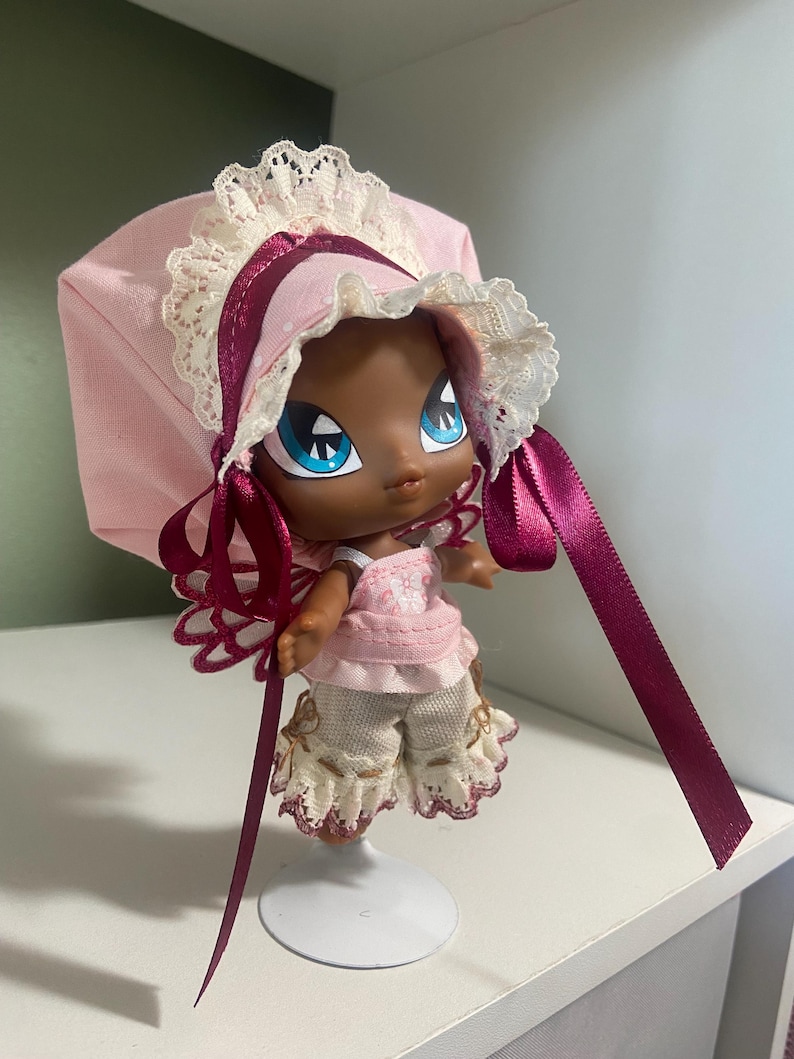 May include: A doll with dark skin, large blue eyes, and a pink bonnet with lace trim and burgundy ribbons. The doll wears a pink dress and cream-colored shorts with lace accents. It stands on a white metal stand.