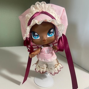 May include: A doll with dark skin, large blue eyes, and a pink outfit. The doll wears a pink bonnet with white lace trim and burgundy ribbons. The doll's shorts are cream-colored with a scalloped edge.