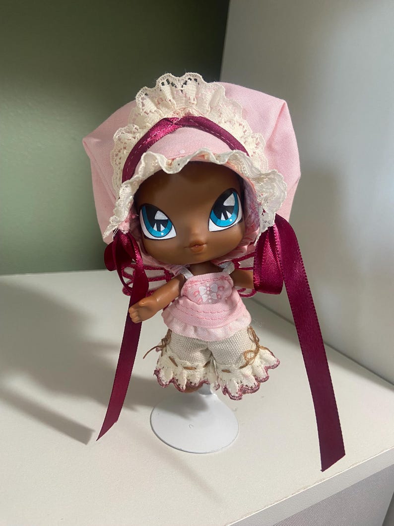 May include: A doll with dark skin, large blue eyes, and a pink outfit. The doll wears a pink bonnet with white lace trim and burgundy ribbons. The doll's shorts are cream-colored with a scalloped edge.