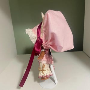 May include: A small doll wearing a pink bonnet with a matching dress and white lace trim. The doll has a burgundy ribbon and is standing on a white metal stand. The doll's legs are visible, with white lace-trimmed pants.