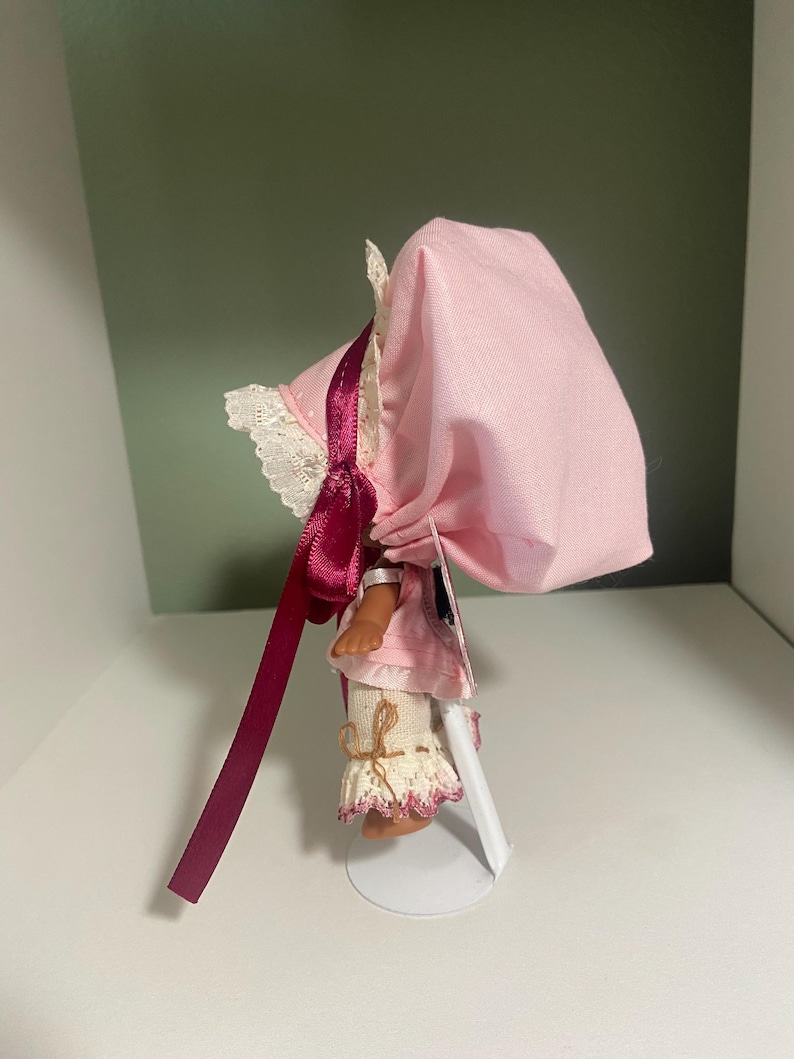 May include: A small doll wearing a pink bonnet with a matching dress and white lace trim. The doll has a burgundy ribbon and is standing on a white metal stand. The doll's legs are visible, with white lace-trimmed pants.