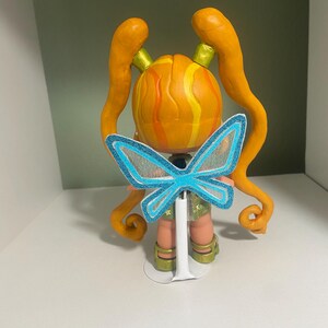 Op de afbeelding: Handgemaakte figuur van een personage met oranje haar gestyled in twee lange staarten. De figuur heeft een ronde kop met gele en oranje strepen. Het draagt een lichtgroene korte broek en heeft blauwe vlindervleugels.