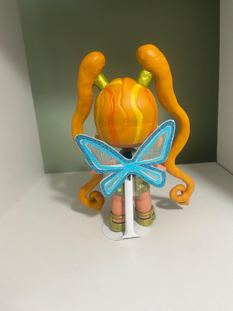 Op de afbeelding: Handgemaakte figuur van een personage met oranje haar gestyled in twee lange staarten. De figuur heeft een ronde kop met gele en oranje strepen. Het draagt een lichtgroene korte broek en heeft blauwe vlindervleugels.