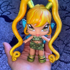 Op de afbeelding: Een pop met een fee&euml;nthema, met een gele en oranje haarstijl en lange krullen. De pop draagt een groene outfit met gouden accenten, blauwe glittervleugels en gouden plateauschoenen. De pop wordt in een hand gehouden.
