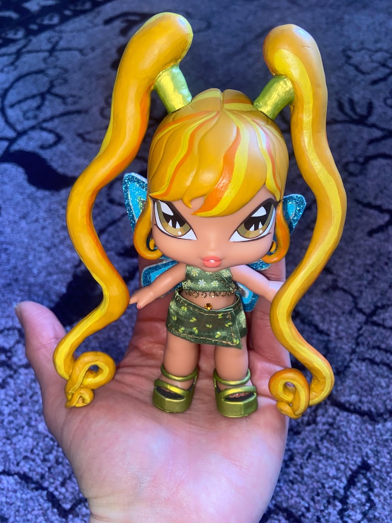 Op de afbeelding: Een pop met een fee&euml;nthema, met een gele en oranje haarstijl en lange krullen. De pop draagt een groene outfit met gouden accenten, blauwe glittervleugels en gouden plateauschoenen. De pop wordt in een hand gehouden.