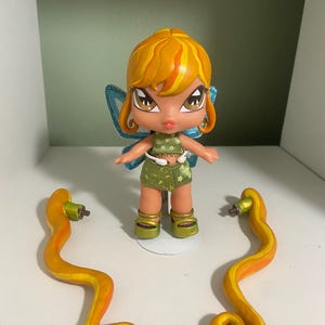 Op de afbeelding: Een Bratz-achtige pop met oranje haar, een groen en witte outfit en blauwe vleugels. Twee oranje en gele slangachtige accessoires zijn aan weerszijden van de pop geplaatst, alles op een witte plank.