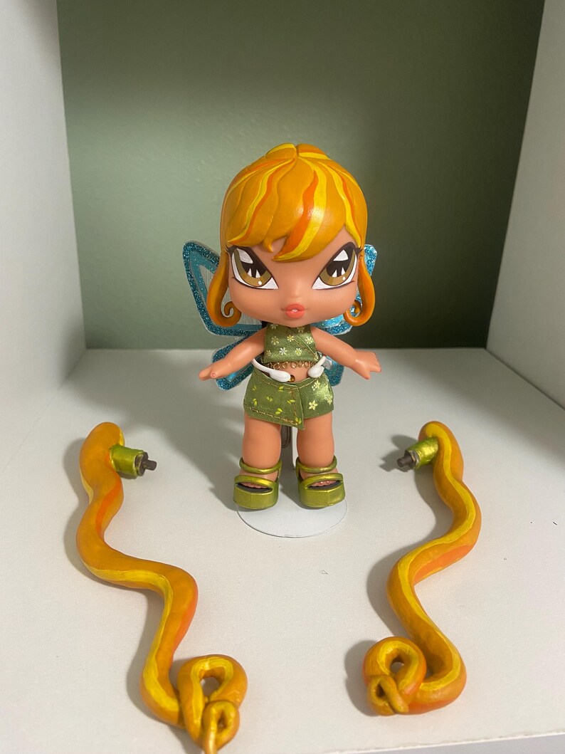 Op de afbeelding: Een Bratz-achtige pop met oranje haar, een groen en witte outfit en blauwe vleugels. Twee oranje en gele slangachtige accessoires zijn aan weerszijden van de pop geplaatst, alles op een witte plank.