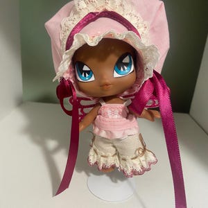 May include: A doll with a dark complexion, large blue eyes, and a pink bonnet with white lace trim. The doll wears a pink top and cream-colored shorts with embroidered details. Long, dark pink ribbons hang from the bonnet.