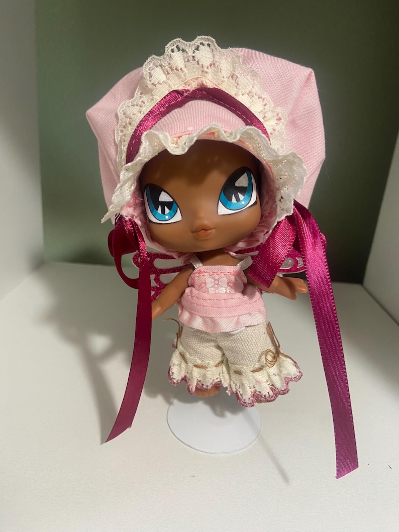 May include: A doll with a dark complexion, large blue eyes, and a pink bonnet with white lace trim. The doll wears a pink top and cream-colored shorts with embroidered details. Long, dark pink ribbons hang from the bonnet.