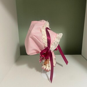 May include: A pink bonnet with white lace trim and a burgundy ribbon. The bonnet is displayed on a white stand against a white shelf and a green wall. The bonnet has a vintage or antique aesthetic.