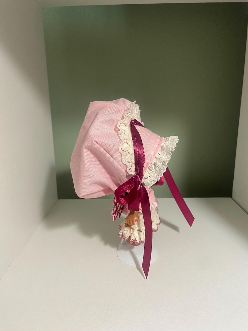 May include: A pink bonnet with white lace trim and a burgundy ribbon. The bonnet is displayed on a white stand against a white shelf and a green wall. The bonnet has a vintage or antique aesthetic.