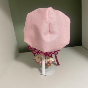 May include: A light pink bonnet with a ruffled trim and a maroon ribbon. The bonnet is displayed on a white stand. The bonnet is made of a soft fabric and is likely for a baby or doll.