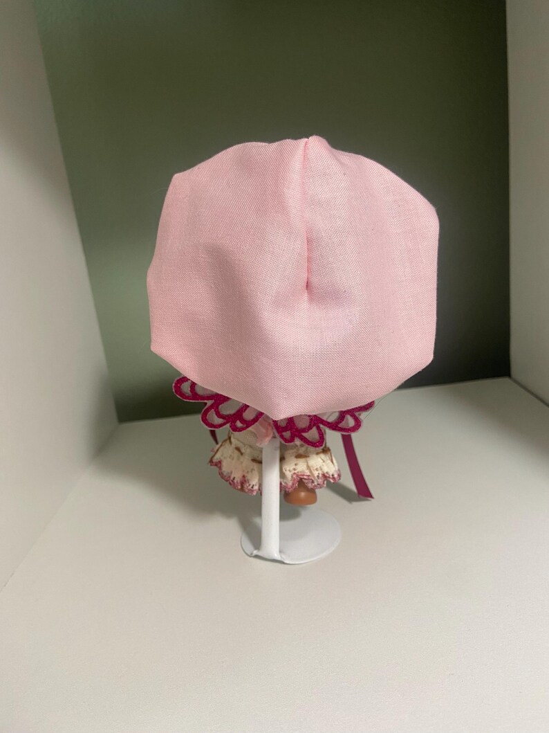 May include: A light pink bonnet with a ruffled trim and a maroon ribbon. The bonnet is displayed on a white stand. The bonnet is made of a soft fabric and is likely for a baby or doll.