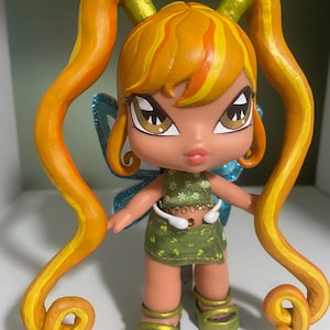 Op de afbeelding: Een aangepaste pop met oranje en geel haar in staartjes. De pop heeft bruine ogen, een groene outfit en groene plateausandalen. De pop heeft blauwe glittervleugels en staat op een wit oppervlak.