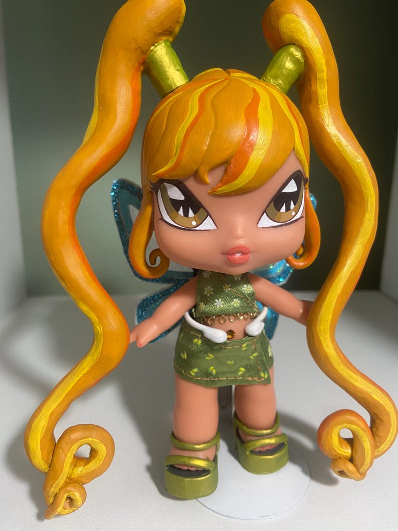 Op de afbeelding: Een aangepaste pop met oranje en geel haar in staartjes. De pop heeft bruine ogen, een groene outfit en groene plateausandalen. De pop heeft blauwe glittervleugels en staat op een wit oppervlak.