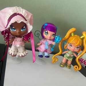 May include: Three Bratz-style fairy dolls with large eyes and stylized features. One doll wears a pink bonnet and outfit, another has purple hair and a blue wand, and the third has orange pigtails and a green dress. Each doll has wings.