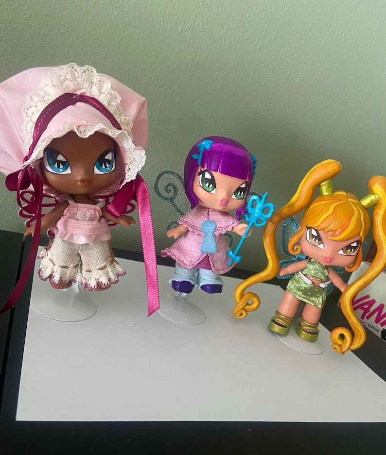 May include: Three Bratz-style fairy dolls with large eyes and stylized features. One doll wears a pink bonnet and outfit, another has purple hair and a blue wand, and the third has orange pigtails and a green dress. Each doll has wings.