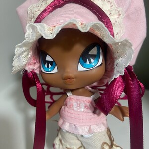 May include: A doll with dark skin, large blue eyes, and a pink bonnet with white lace trim. The doll wears a pink top and cream-colored shorts with lace. Burgundy ribbons are tied on the bonnet. The doll has a whimsical, cartoonish appearance.