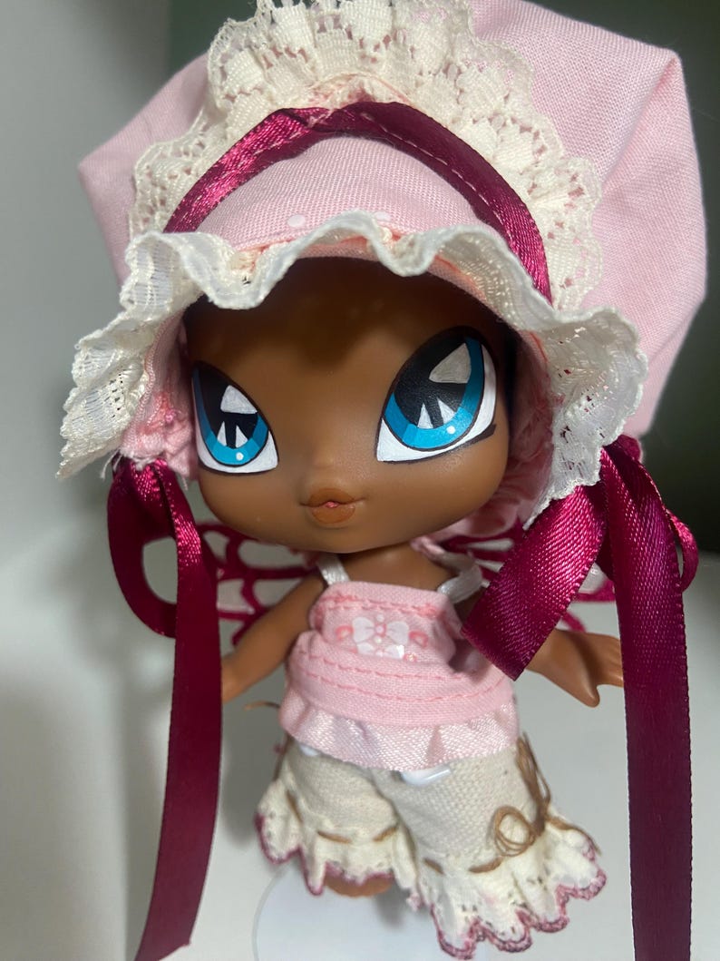 May include: A doll with dark skin, large blue eyes, and a pink bonnet with white lace trim. The doll wears a pink top and cream-colored shorts with lace. Burgundy ribbons are tied on the bonnet. The doll has a whimsical, cartoonish appearance.