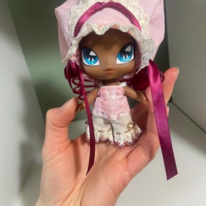May include: A small doll with dark skin, large blue eyes, and a pink bonnet with lace trim. The doll wears a pink top and white pants with lace details. Long, dark pink ribbons hang from the bonnet.