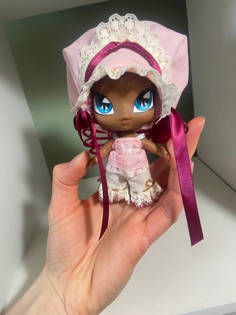May include: A small doll with dark skin, large blue eyes, and a pink bonnet with lace trim. The doll wears a pink top and white pants with lace details. Long, dark pink ribbons hang from the bonnet.
