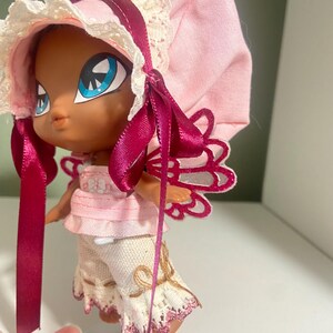 May include: A doll with a dark complexion, large blue eyes, and a pink bonnet with lace trim. The doll is wearing a pink top, white pants with lace trim, and has pink and white wings. A dark pink ribbon is tied around the doll's neck and bonnet.