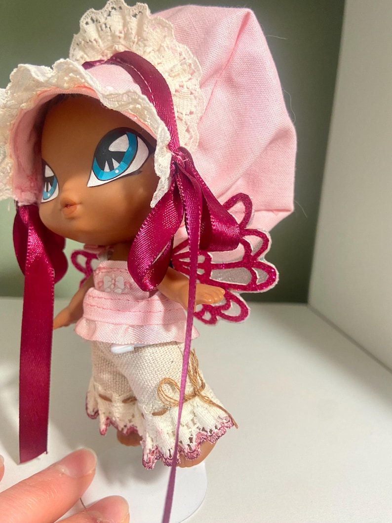 May include: A doll with a dark complexion, large blue eyes, and a pink bonnet with lace trim. The doll is wearing a pink top, white pants with lace trim, and has pink and white wings. A dark pink ribbon is tied around the doll's neck and bonnet.