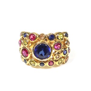 May include: A wide gold band ring featuring a large central round blue gemstone surrounded by a cluster of smaller, multi-colored round stones including pink, yellow, and blue gems set in individual gold bezels across the entire surface.