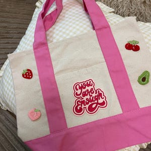 May include: A cream-colored canvas tote bag with bright pink handles and a matching pink base. The bag features embroidered patches of a strawberry, a peach, cherries, and an avocado, along with a central patch that reads You are Enough.