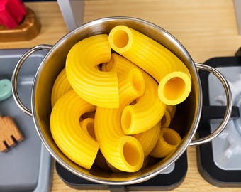May include: A stainless steel pot filled with several bright yellow, curved macaroni pasta shapes. The pot features two side handles and is positioned on a stovetop surface. The pasta pieces are hollow, tubular, and have a smooth texture.