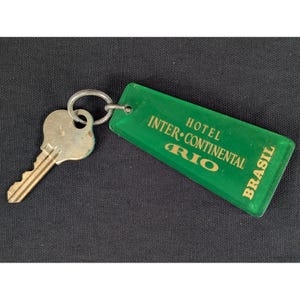 May include: A silver-toned metal key attached to a translucent green rectangular plastic hotel keychain. The keychain features gold-colored text that reads HOTEL INTER-CONTINENTAL RIO BRASIL, with the word RIO appearing in a larger font size.