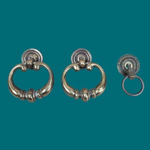 Può includere: Tre maniglie per mobili in ottone composte da due maniglie a goccia più grandi e ornate con bracci curvi decorativi e una piastra di montaggio circolare, insieme a una maniglia ad anello più piccola e semplice con una piastra smerlata.