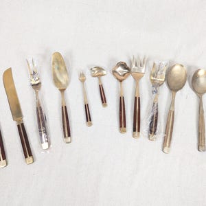 May include: A selection of vintage cutlery, including knives, forks, spoons, and serving utensils. The cutlery has gold-toned metal with dark brown wooden handles. The arrangement is on a white surface, displaying the different pieces.