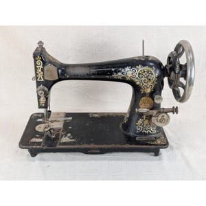 May include: A vintage black sewing machine featuring intricate gold and floral decals along the arm and base. The machine includes a large metal handwheel on the right side, a needle bar, and a flat rectangular base with visible wear and patina.