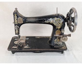 May include: A vintage black sewing machine featuring intricate gold and floral decals along the arm and base. The machine includes a large metal handwheel on the right side, a needle bar, and a flat rectangular base with visible wear and patina.