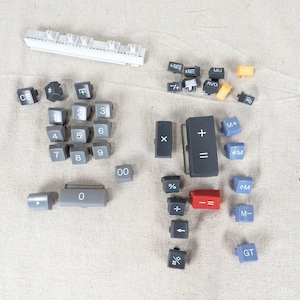 May include: A collection of calculator keys in various colors and sizes, including white, gray, red, and blue. The keys are scattered on a light-colored surface. Some keys have numbers, while others have symbols and functions like "+", "-", and "GT".